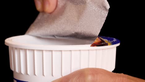 a hand peels back the foil lid of an instant noodle cup under bright studio lighting, captured in a close-up with a black background and smooth camera movement