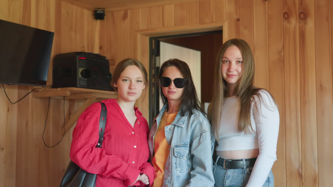 Three young women stand indoors against wooden wall, posing together with calm expressions as woman in middle lifts sunglasses, surrounded by casual setting with visible speaker and television
