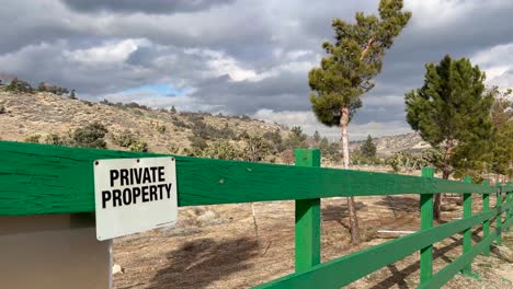 Cartel-De-Propiedad-Privada-En-Una-Valla-Verde-En-El-Desierto-Alto-Con-árboles-Que-Soplan-En-El-Viento-Y-Un-Cielo-Nublado-Y-Cambiante