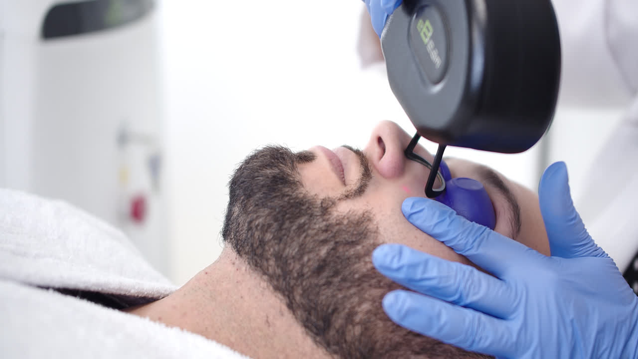Beautiful slow motion profile shot of a handsome young man receiving a beauty treatment with a laser device on his face by a female doctor wearing gloves.