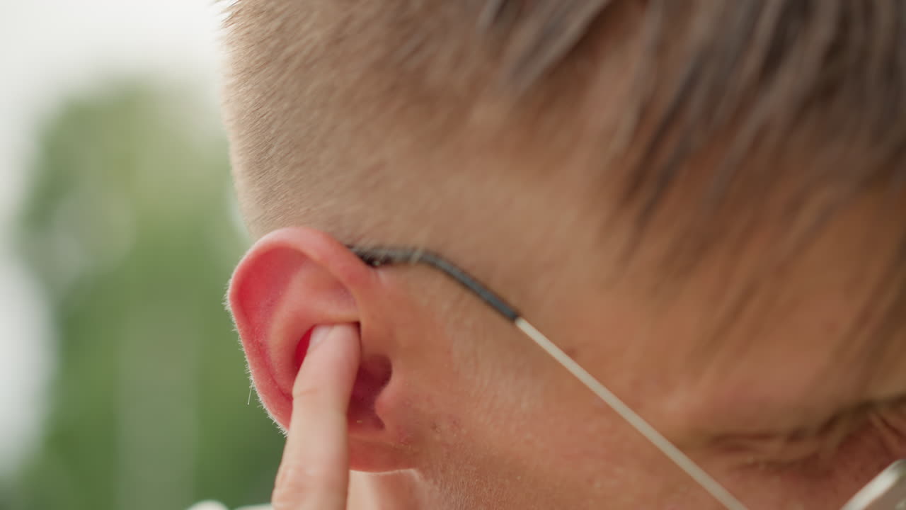 Primer plano de un hombre caucásico quitándose un auricular y frotándose la oreja, gotas de lluvia y sudor en la piel, carril del parque de fondo, corredor deteniéndose para ajustar los auriculares, viajero molesto por el sonido, detalle urbano espontáneo