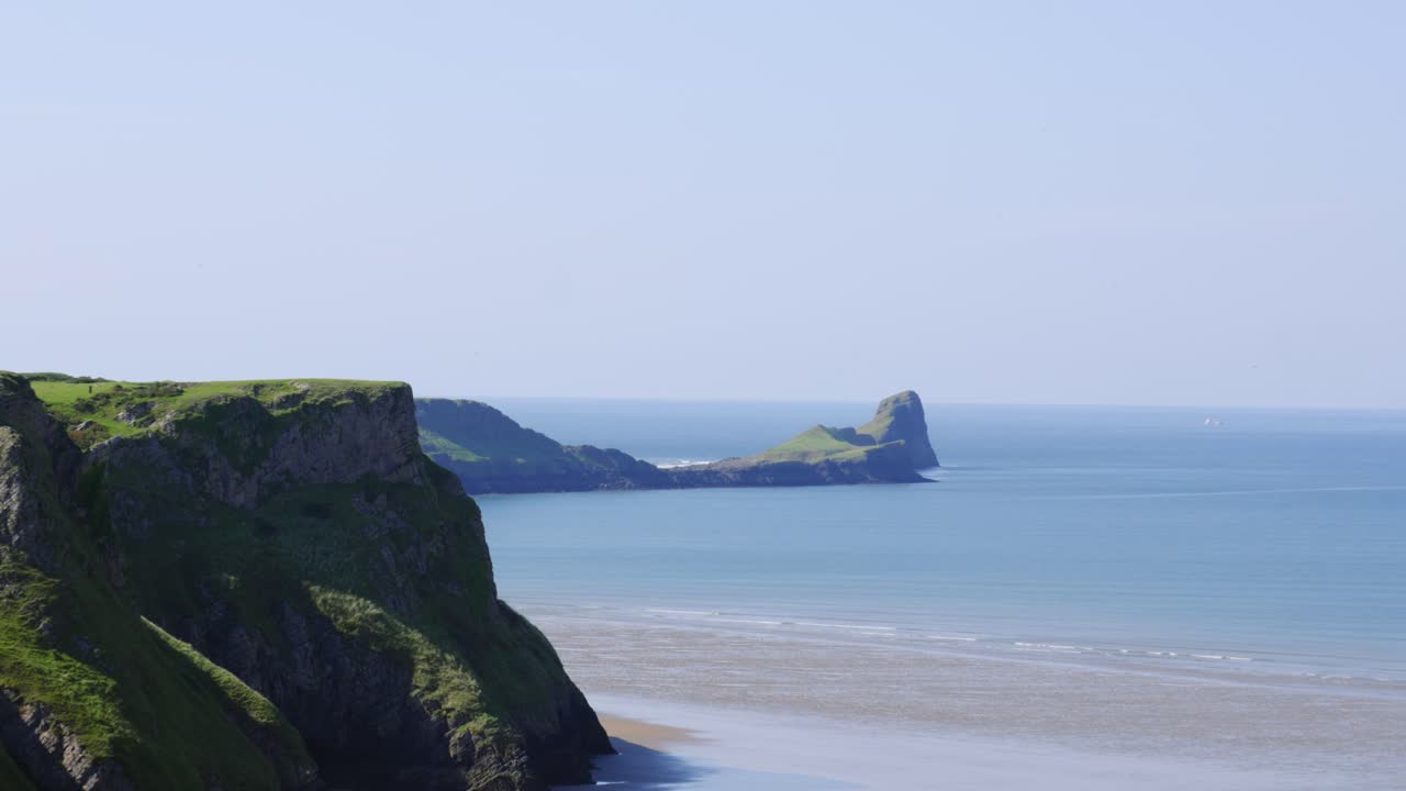 Pan from Path to Rhossili Bay Across Steep Cliffs with Tourist Silhouettes and Worm's Head Landmark with Small Waves 4K