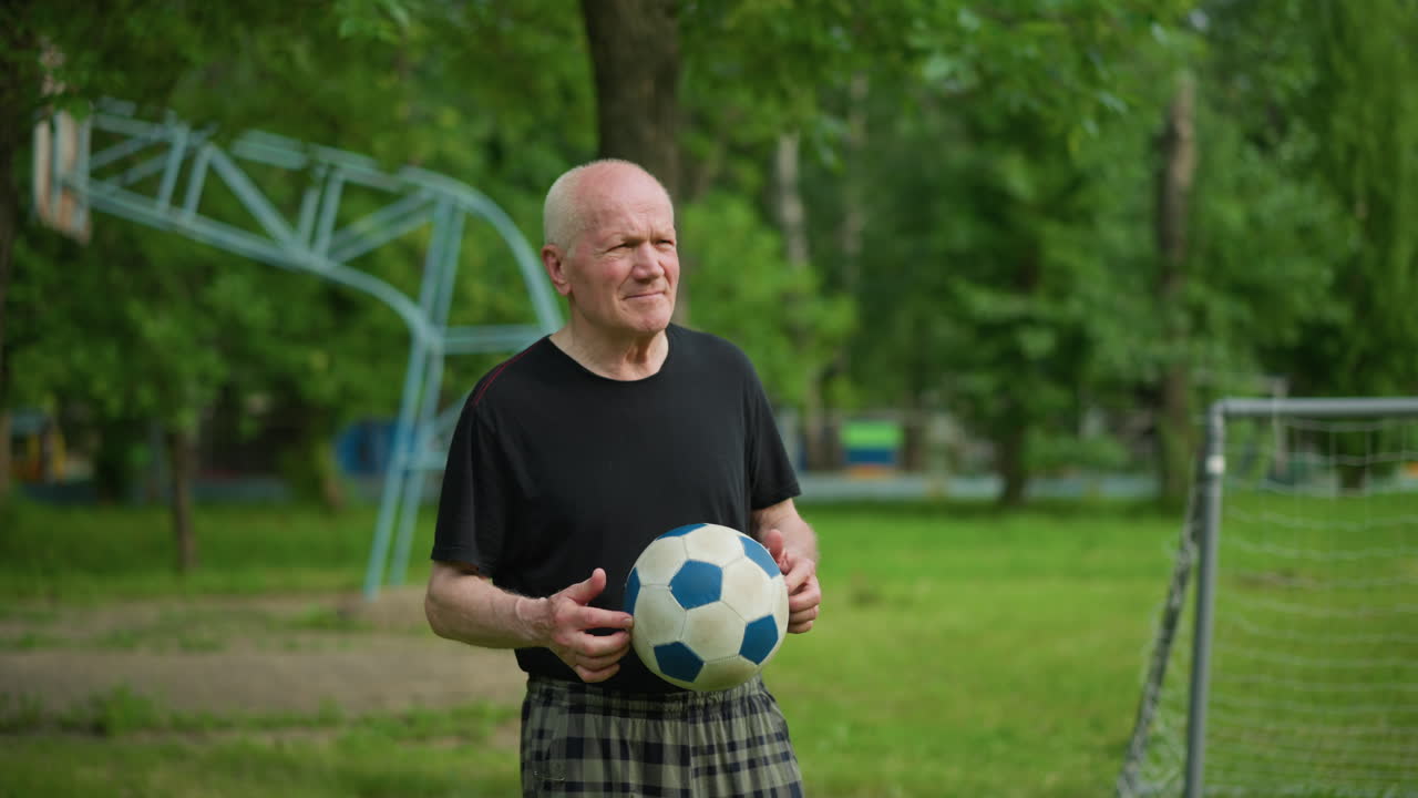 un hombre mayor, centrado y contemplativo, rodando una pelota de fútbol en la mano mientras mira hacia abajo, con una vista de fondo de poste de gol y equipos al aire libre en el fondo