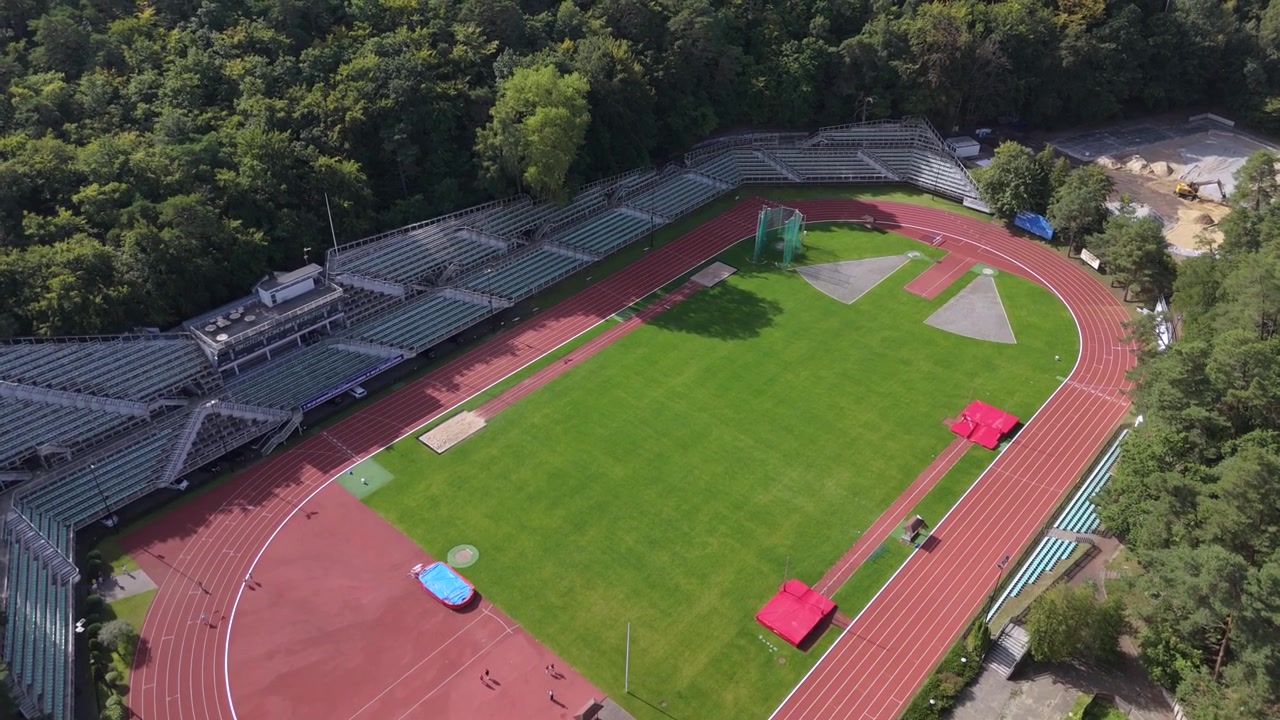 Vista aérea vertical en ángulo picado de una pista de atletismo profesional, un campo verde y las gradas de un estadio junto a un bosque, destacando la salud, el entrenamiento deportivo y un estilo de vida activo