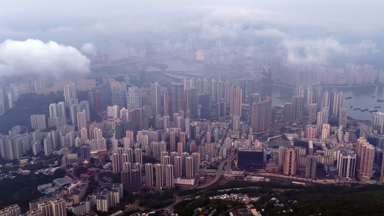 Aerial view of dense Hong Kong cityscape under foggy twilight, featuring high-rise buildings, residential towers, harbor, and mountains in the background