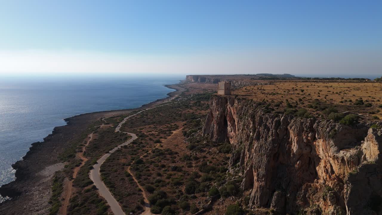 Zoom in con ripresa aerea che mostra una strada costiera tortuosa lungo la costa siciliana, con scogliere rocciose e mare blu intenso sotto la luce del giorno