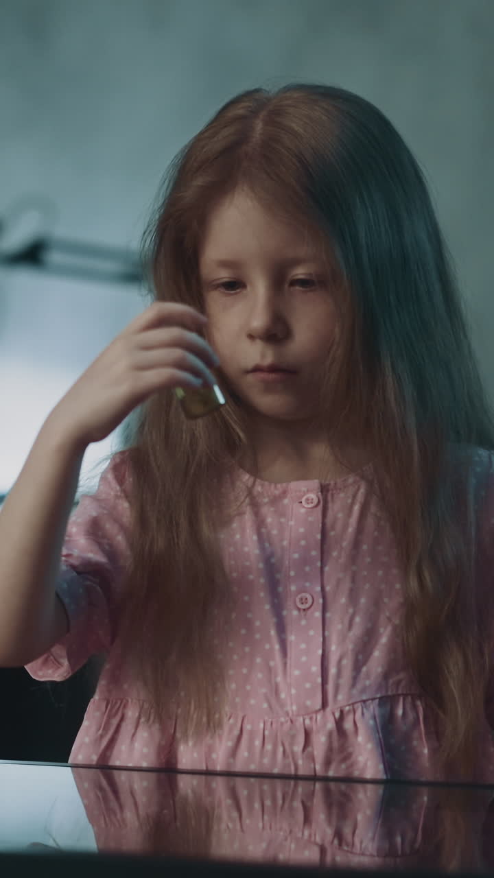 Red long haired little girl takes pill bottle of small vitamin medicines to examine sitting at glass table in semi-dark room at home close view slow motion