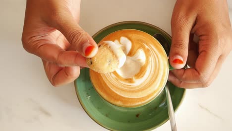 person holding a coffee cup with latte art and dipping a cookie in it