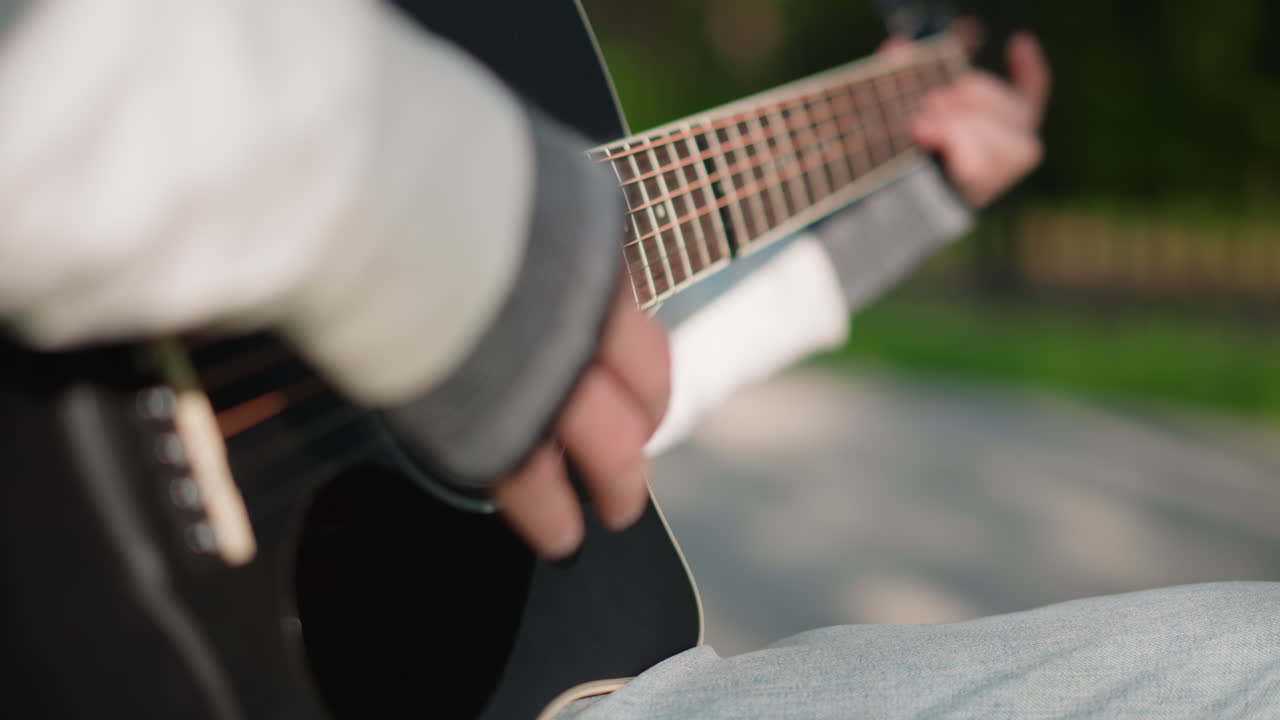 closeup guitarist playing acoustic guitar, gentle fingerstyle on black body, denim sleeve, park pathway, warm sunlight, reflective songwriter practice, intimate lofi vibe, shallow depth, soft bokeh