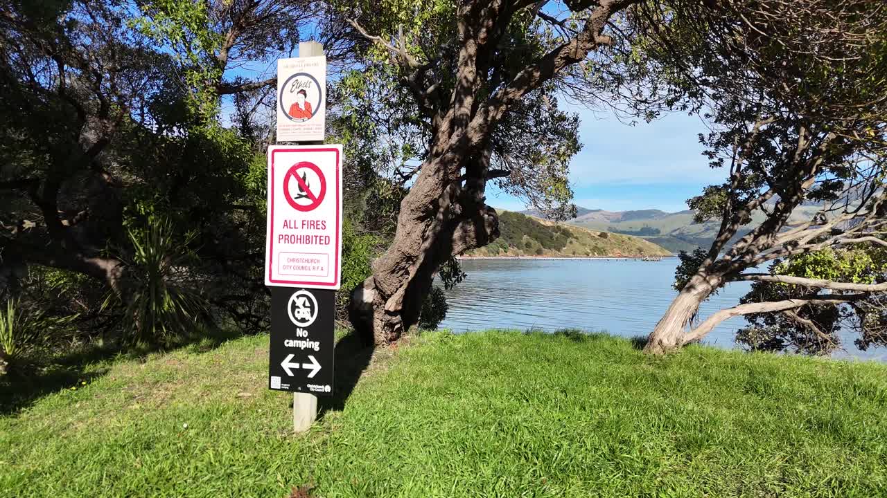 A serene lakeside scene in Akaroa with a prominent warning sign amidst lush greenery and clear skies
