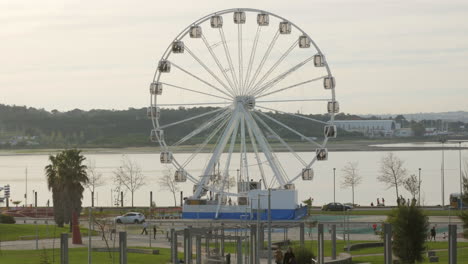 people riding ferris wheel with glass cabins spinning in an amusement park by the riverside - wide shot