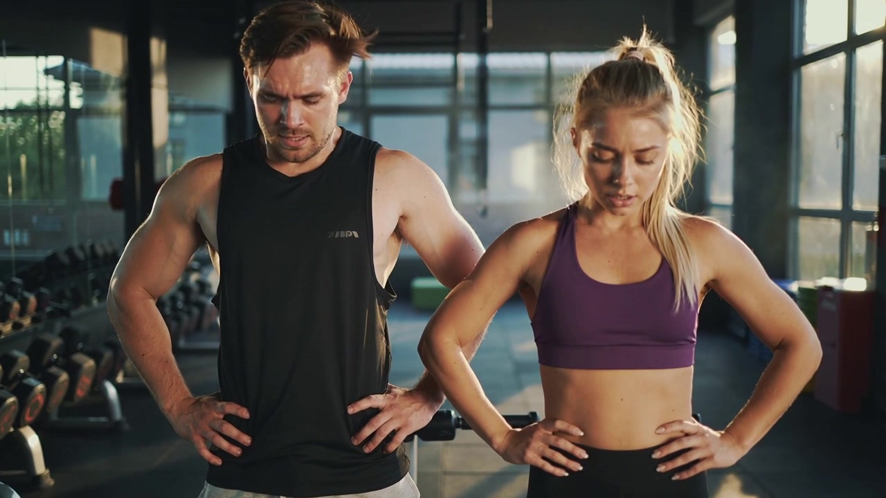 Nach dem Satz verschnaufend, Mann in schwarzem Oberteil und Frau in Lila stehen im Fitnessstudio mit Hanteln