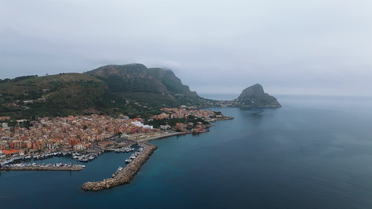 Drone footage showing Porticello’s harbor and Capo Zafferano headland near Palermo, Sicily, framed by mountains, dramatic coastline and calm Mediterranean waters