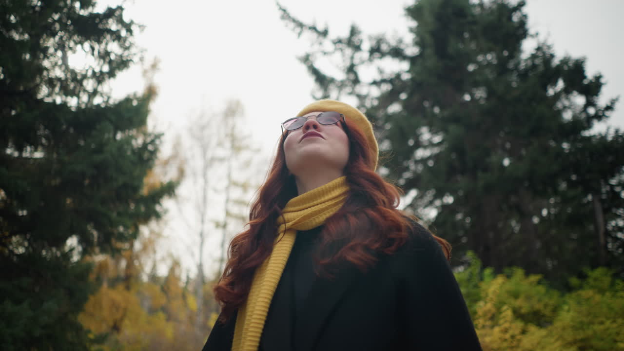 una dama universitaria de cabello rojo fluido mira hacia arriba, vestida con un abrigo negro, boina amarilla y una bufanda a juego, vagando por un exuberante parque adornado con colores de otoño