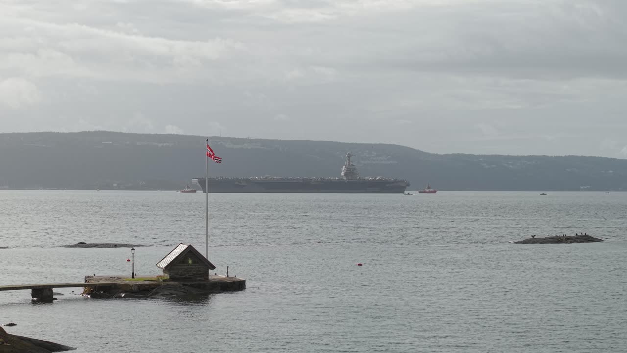 Island With A Flagpole And Hut With USS Gerald R. Ford Aircraft Carrier Cruising In The Background In Oslo, Norway. - wide shot
