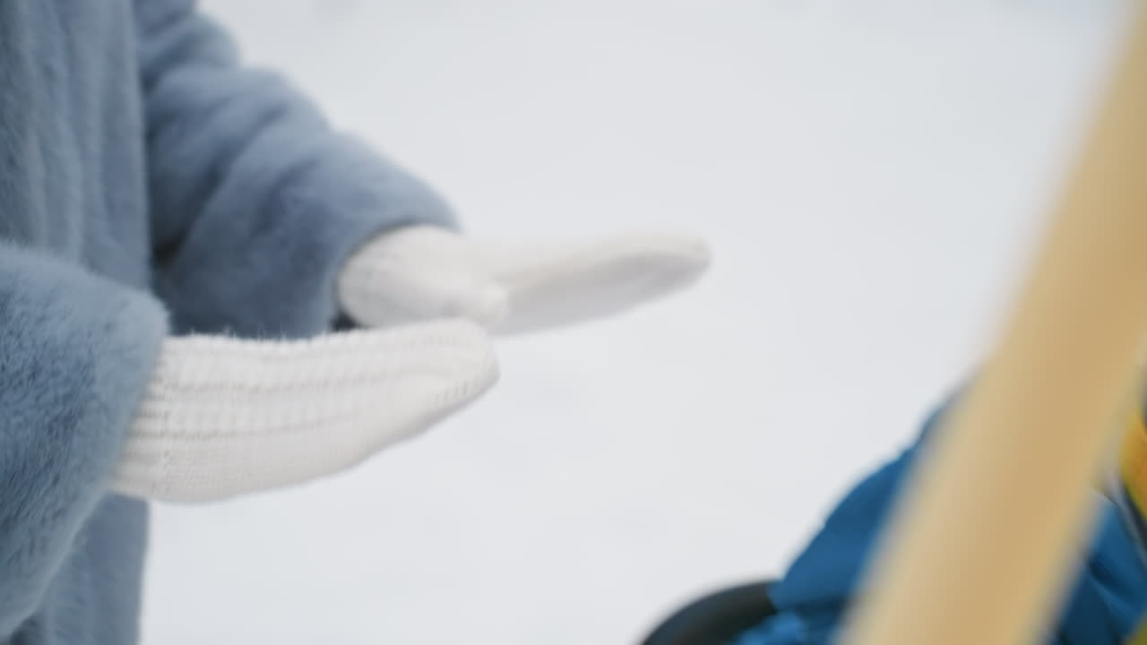 雪の中の母と息子の手。青いスノースーツを着た子供に暖かさを与える白いミトンがクローズアップされている。優しい手の触れ合い、居心地の良い毛皮のコート、静かな公園、柔らかな冬の光、そして映画のような浅い被写界深度が、優しさを際立たせる。