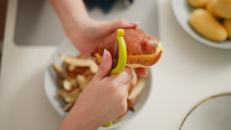 close up hand view of person peeling potato over bowl filled with potato skins, showing detailed focus on kitchen task in well-lit cooking environment