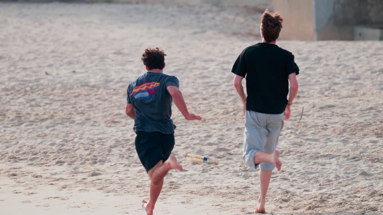 Two young men run barefoot across the sand on a sunny day, enjoying outdoor exercise and friendship