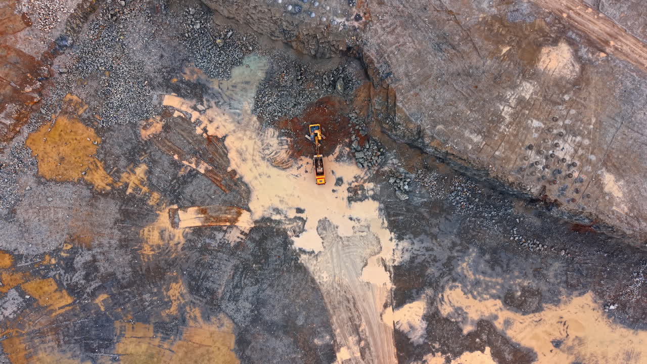 Heavy machinery works in a mining site. Two large machines operate in a mine digging and moving soil and rocks. The area shows different colors and textures