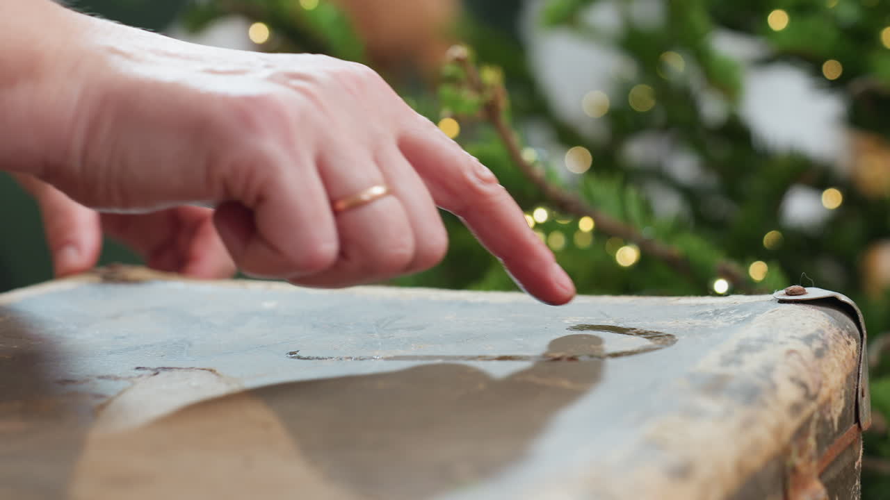 Close hand view of hand sketching shapes on dusty box surface using wet finger, capturing tactile creative moment with blurred glowing Christmas tree lights in background during cozy festive