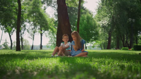 Hermanos-Sonrientes-Juegan-En-La-Hierba-Bajo-Un-árbol.-Niños-Felices-Sentados-En-El-Césped.