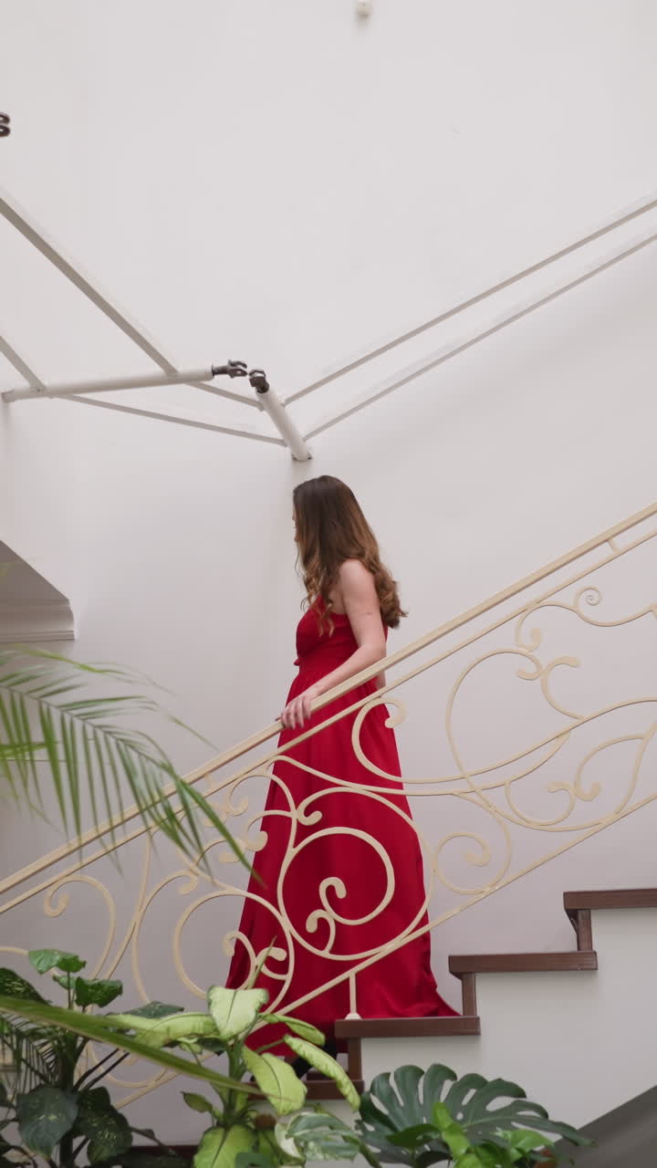 빨간 옷을 입은 예쁜 여자가 계단을 내려간다. 빨간 슬리트 드레스를 입은 갈색 느슨한 머리카락을 가진 아름다운 여자가 고급 주택에서 파티를 방문합니다.