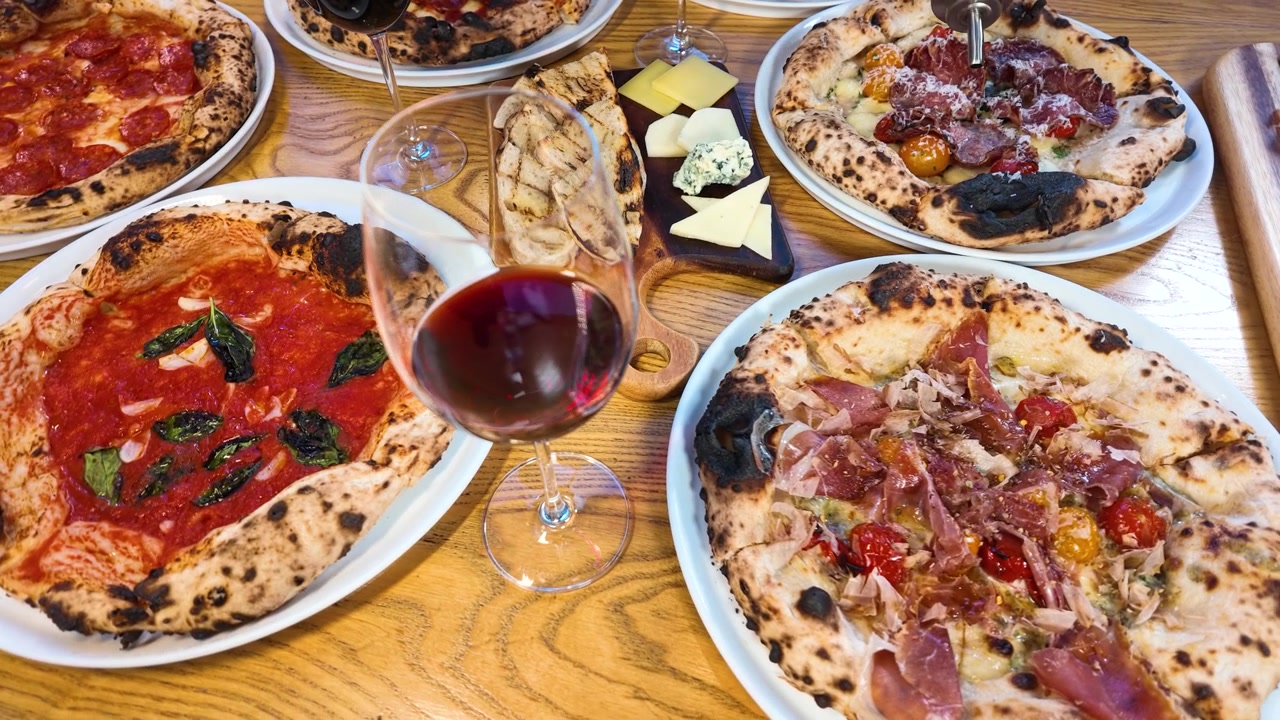 Aufsicht auf mehrere Pizzen, eine Käseplatte und Rotwein auf einem Holztisch.
