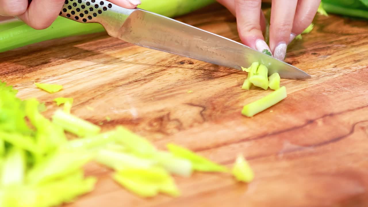 Detailed view of hands slicing celery on a wooden cutting board with a sharp knife.