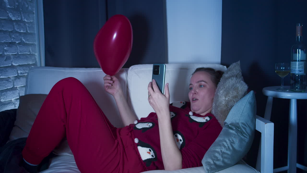 Una giovane donna in abiti casalinghi, sdraiata sul divano, che guarda un video sul suo cellulare che la fa ridere e la stupisce.