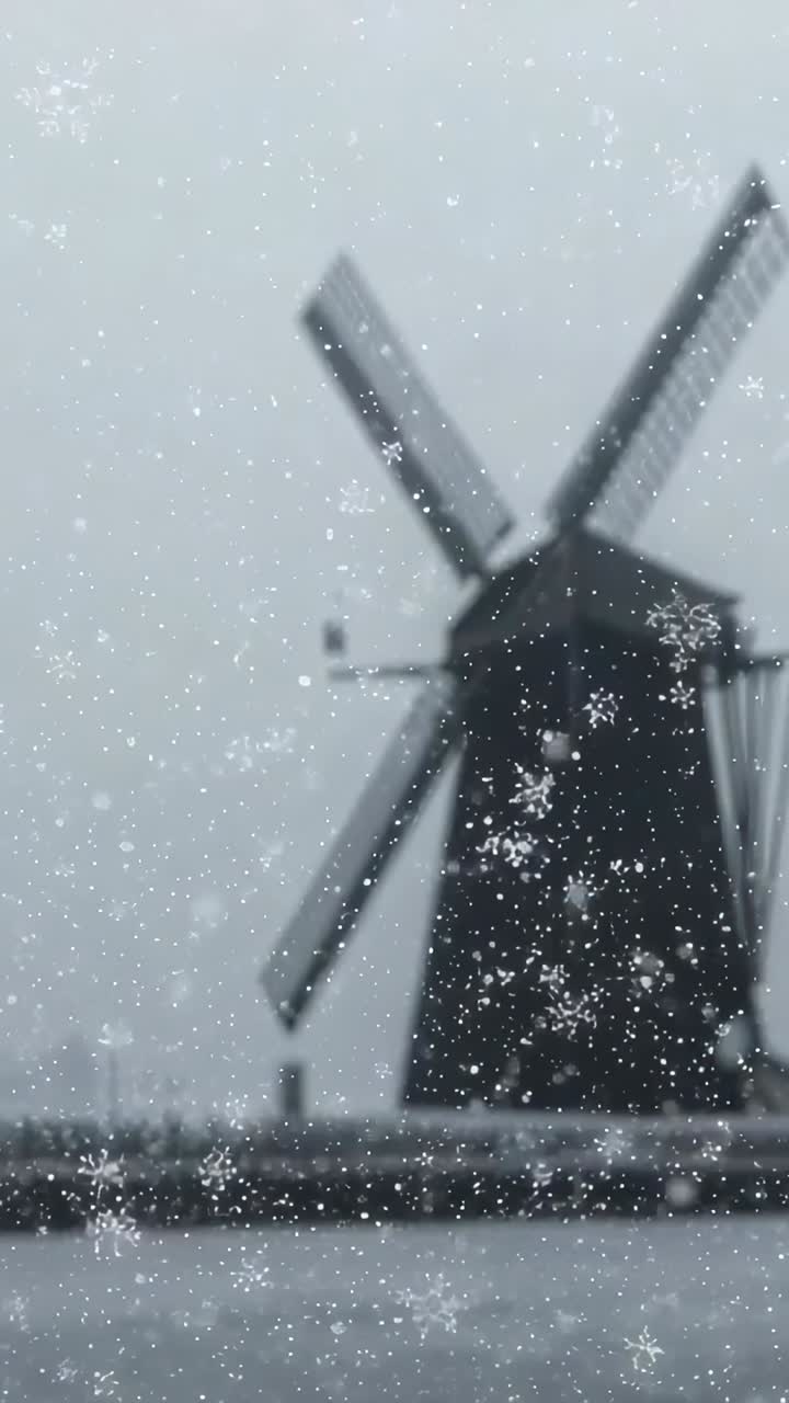 Vertical video: Snow falling then easing, tower-mill turning sails from wind at canal, frozen fence