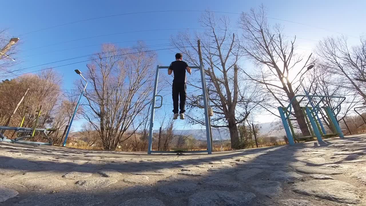 Low wide-angle shot captures a person executing a pull-up on a metal calisthenics bar, surrounded by bare trees and blue sky in a sunlit outdoor fitness area