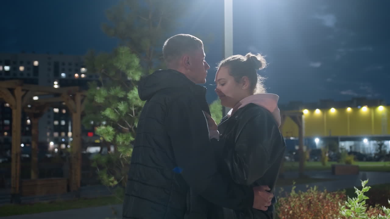 Young man gently kisses wife on forehead under glowing street light at dusk with peaceful expressions and soft shadows on ground surrounded by green park, modern buildings