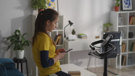 young woman is resting during training with exercise bike at home using smartphone in break time checking social nets internet addiction