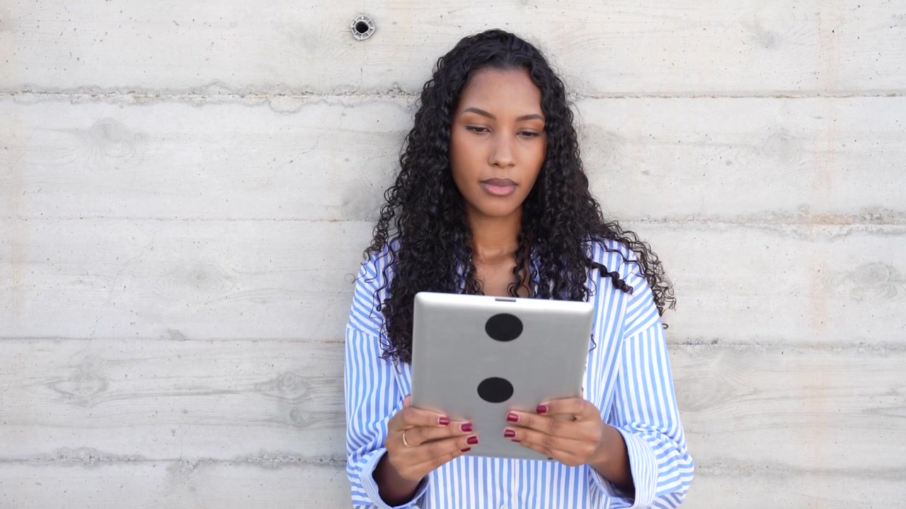 A woman engaged in using a tablet while leaning against a modern concrete wall in the city