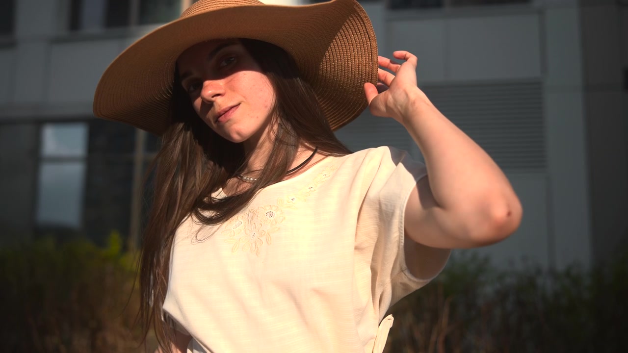 Una giovane donna in un vestito chiaro siede sotto la calda luce del sole su una panchina di legno vicino a piante verdi e un edificio moderno, creando uno stile di vita all'aperto calmo ed elegante