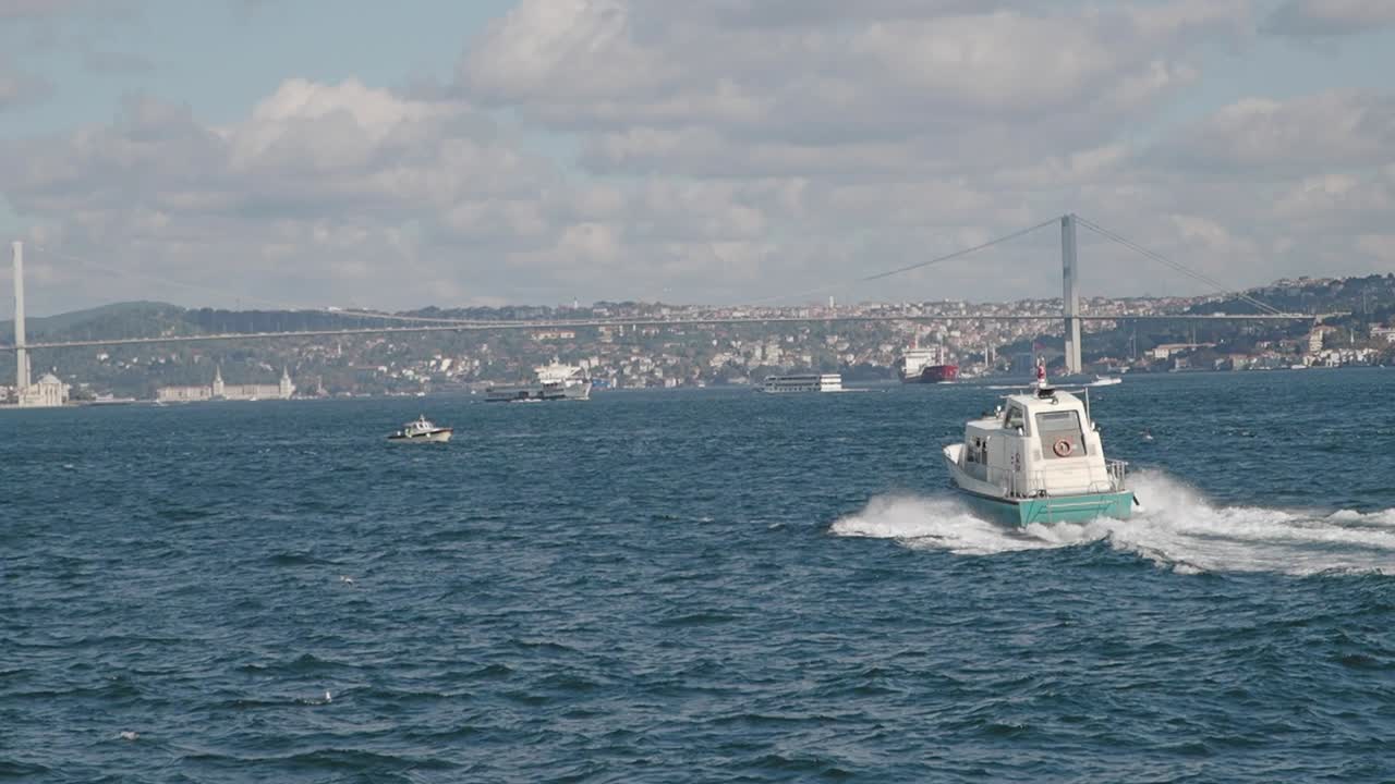 Boats and ships navigating a busy strait with a suspension bridge and cityscape in the background