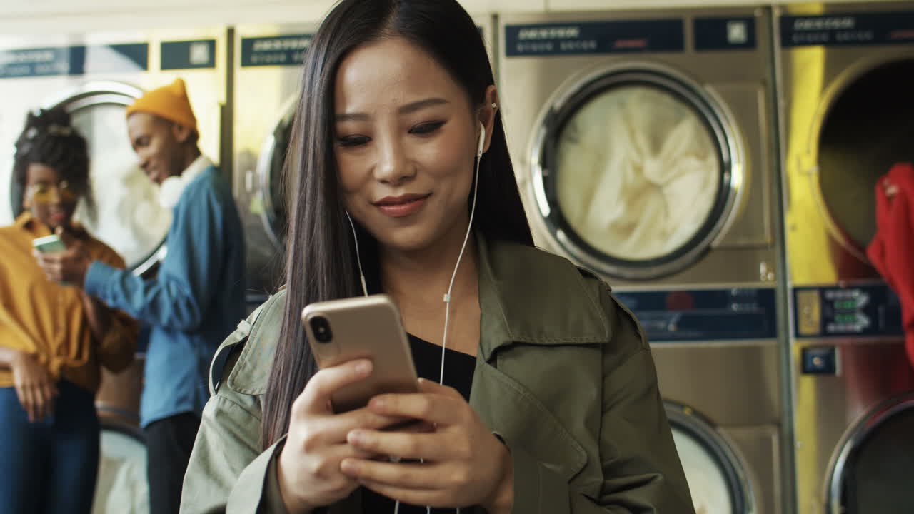 mujer bastante alegre con auriculares viendo videos en un smartphone en la sala de servicio de lavandería