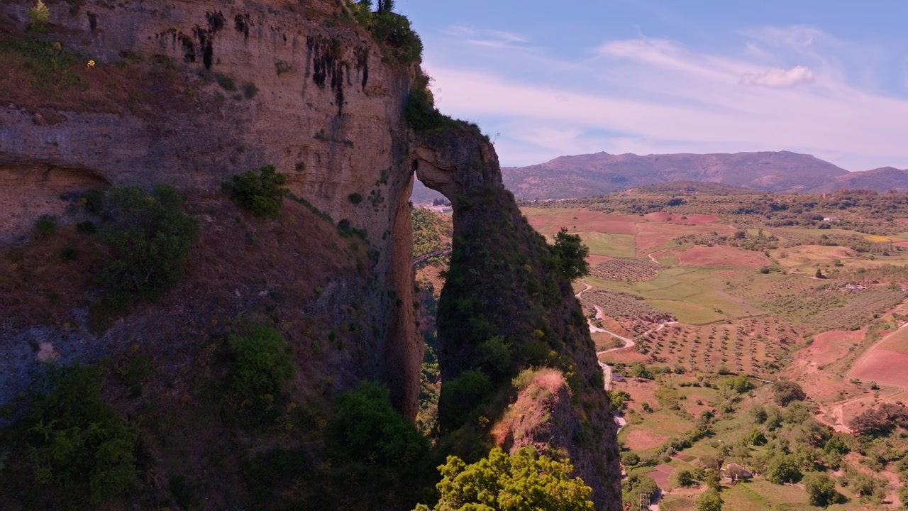 Filmato di drone che scivola lentamente di lato con un grandangolo, passando lungo l'intera parete rocciosa di Ronda, e che mostra la piccola cavità naturale nella roccia incastonata nella vasta parete calcarea e la valle aperta oltre