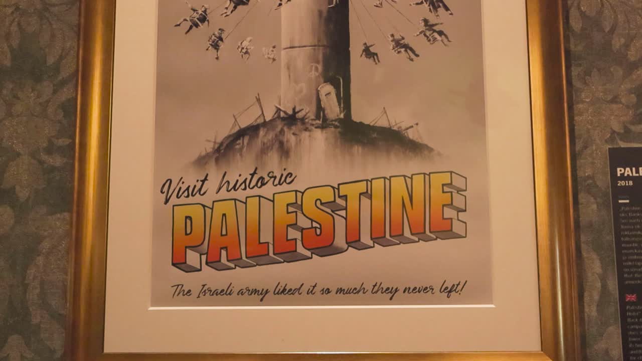 Iconic art made by Banksy called Visit Historic Palestine poster framed in a golden frame on a wall at Banksy exhibition. Shows children using a watch tower as an amusement park while riding on swings