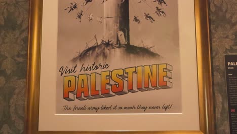 iconic art made by banksy called visit historic palestine poster framed in a golden frame on a wall at banksy exhibition. shows children using a watch tower as an amusement park while riding on swings
