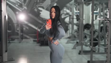 Hermosa-Morena-Curvilínea-Pasa-Tiempo-En-El-Teléfono-Haciendo-Fotos-En-Un-Smartphone-En-El-Espejo-Del-Gimnasio