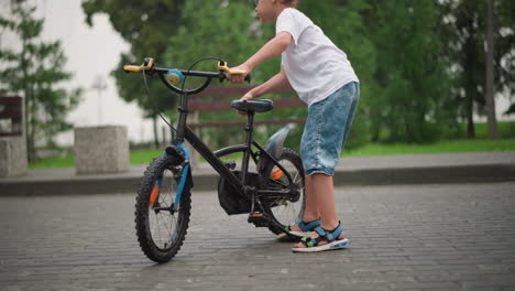 a young boy wearing a white t-shirt and denim shorts is seen adjusting the stand of his black bicycle, trying to park it on a paved path in a park