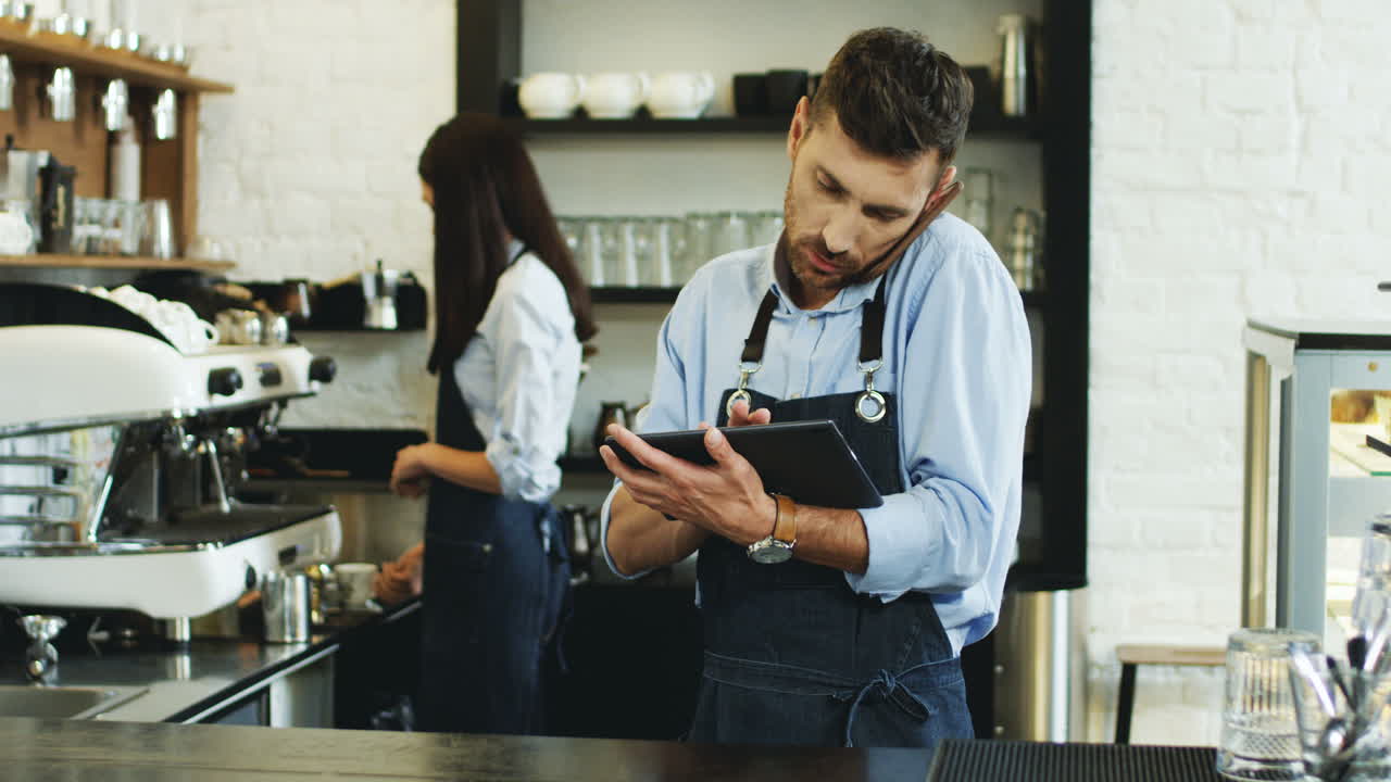 Attractive Waiter Speaking On The Telephone And Using Tablet Computer At The Bar, Brunette Waitress Making Coffee Behind