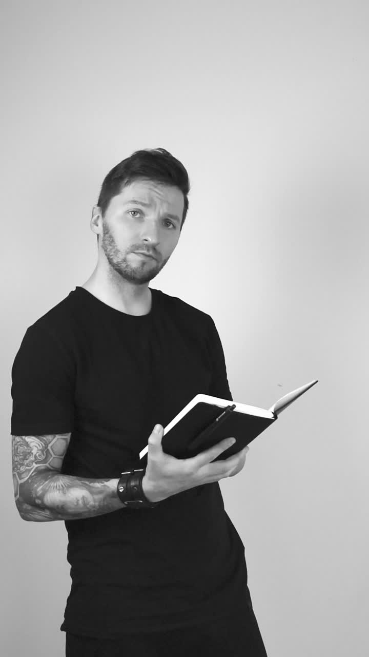 Grayscale footage of a white tattooed man with short hair and beard looking into the camera while holding a notebook or journal, then begins to flip the pages of the little black book he is holding.
