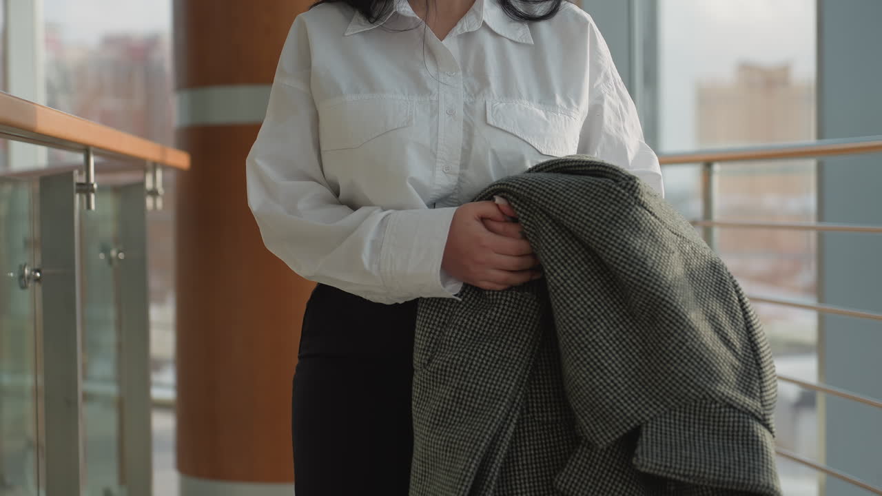 Banker in white shirt holding checkered jacket gazing out through transparent glass frame in bright indoor setting with urban background featuring tall buildings and city street in soft blur