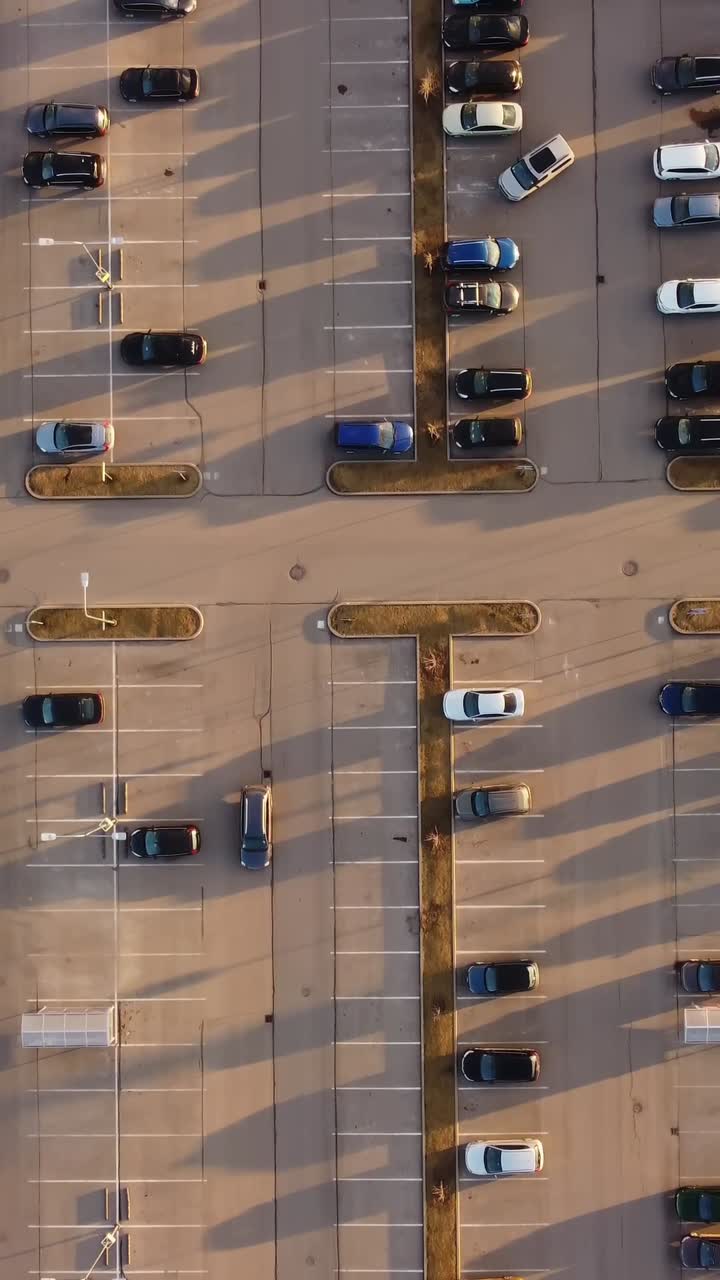 Vertical Aerial view of crowded parking lot during sunset, vehicles navigating tight spaces, elongated shadows stretching across asphalt under golden light