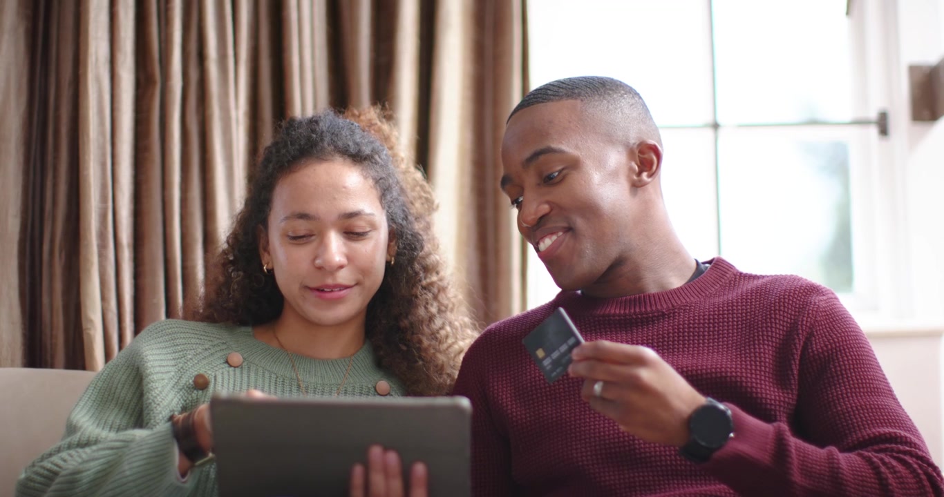 Diverse couple sitting on sofa man holding card up while woman tapping tablet confirming purchase