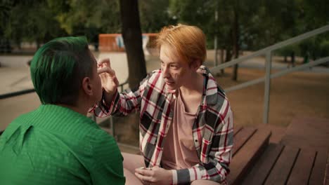 over the shoulder: a girl with short dark yellow hair in a pink checkered shirt puts glitter on the face of her partner a girl with short hair with green hair in a green shirt on brown stands benches near a skate park in a green park in summer