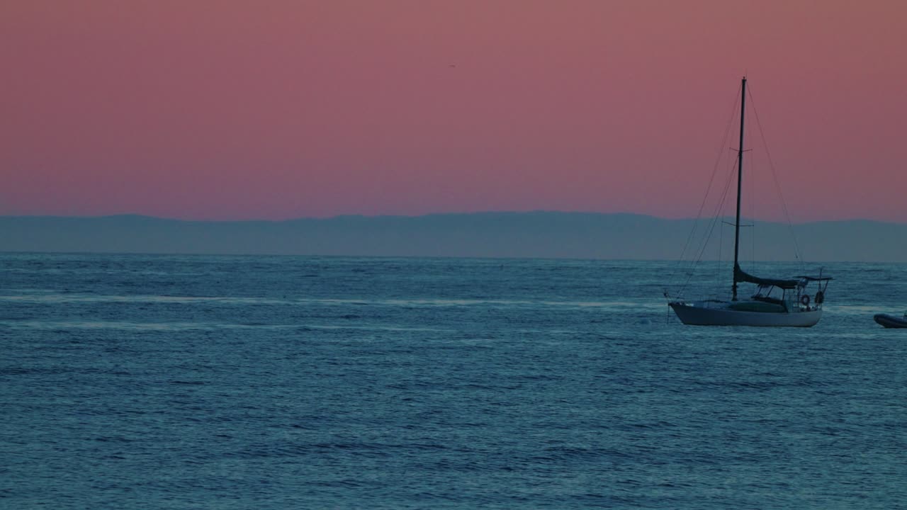 A sailboat rests on calm waters at sunset off the Santa Barbara coast, silhouetted against soft pink skies.