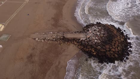 Circling-shot-of-the-water-waves-crashing-the-rocks-where-people-walking-over-the-breakwater-at-the-beach-on-a-mild-climate-day-at-Mar-del-Plata,-Argentina